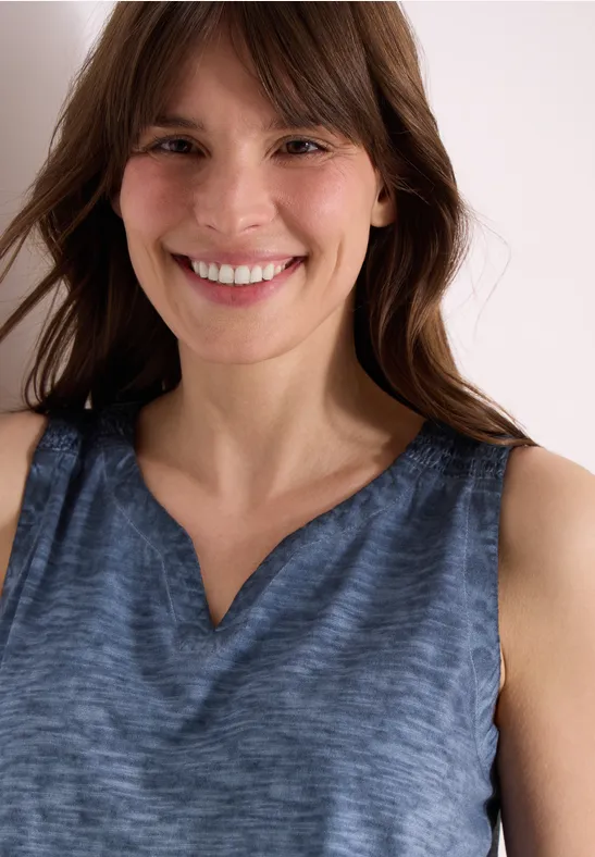 Eine Person trägt ein ärmelloses, blaues Oberteil mit V-Ausschnitt vor hellem Hintergrund.