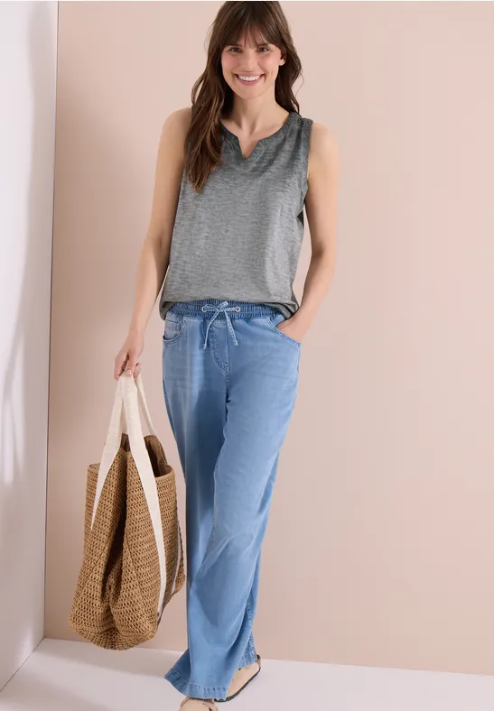 Frau trägt ein graues ärmelloses Top, weite blaue Jeans und eine große geflochtene Tasche