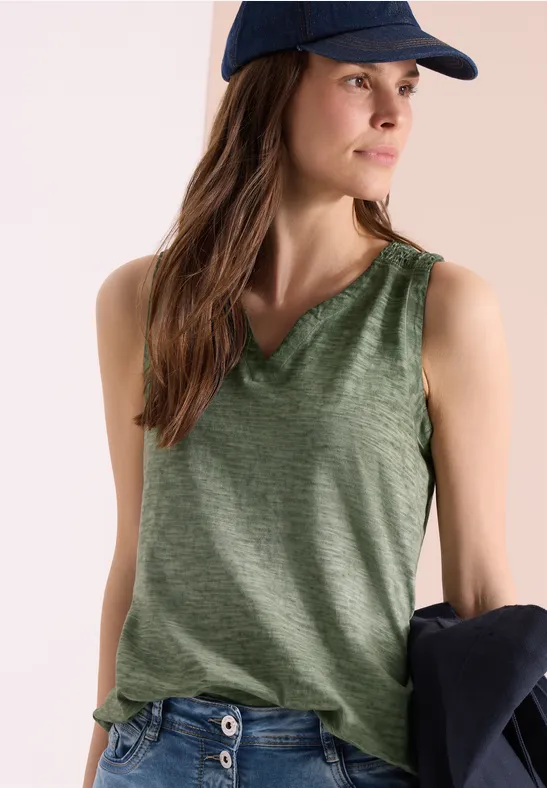 Vrouw in groene mouwloze top en pet, houdt een donkere jas over haar arm.