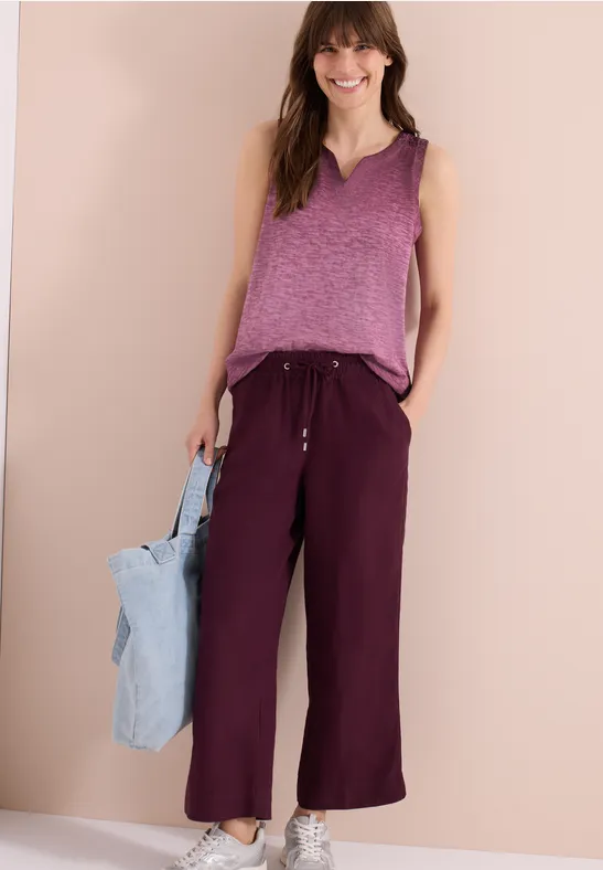 Vrouw in een paarse mouwloze top, wijde wijnrode broek en lichtblauwe tas voor neutrale achtergrond.