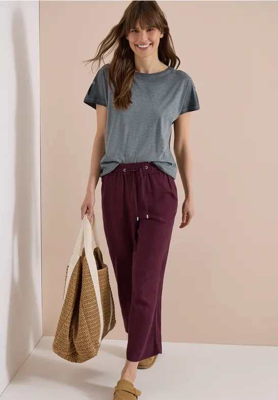 Vrouw in grijs T-shirt en bordeaux wijde broek met gevlochten tas, staand voor een neutrale achtergrond.