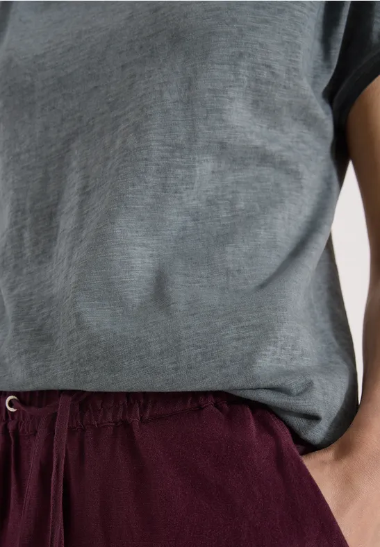 Bovenlichaam met grijs T-shirt en bordeauxrode short, persoon staat ontspannen met hand in zak.