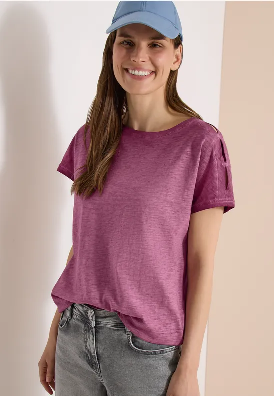 Eine Frau in lässigem Outfit: violettes T-Shirt, helle Jeans und blaue Cap vor hellem Hintergrund.