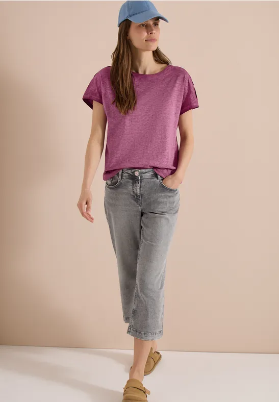 Person trägt lila T-Shirt, graue Jeans, grüne Schuhe und blaue Kappe vor beigem Hintergrund.