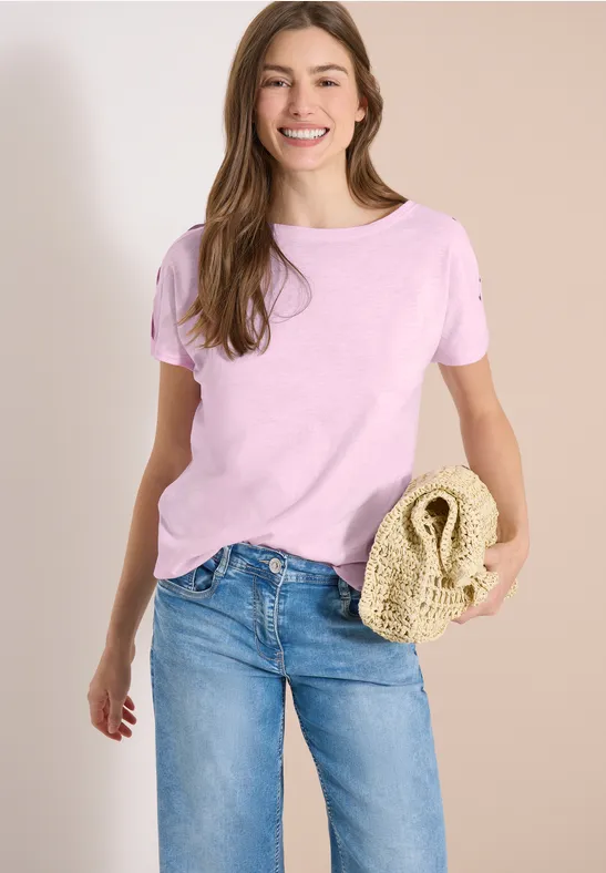 Frau trägt rosa T-Shirt, blaue Jeans und hält eine geflochtene Strohtasche vor hellem Hintergrund.