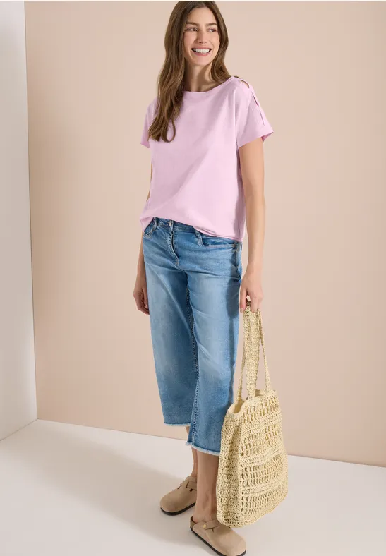 Eine Frau trägt ein rosafarbenes T-Shirt, weite Jeans, Basttasche und flache Schuhe.