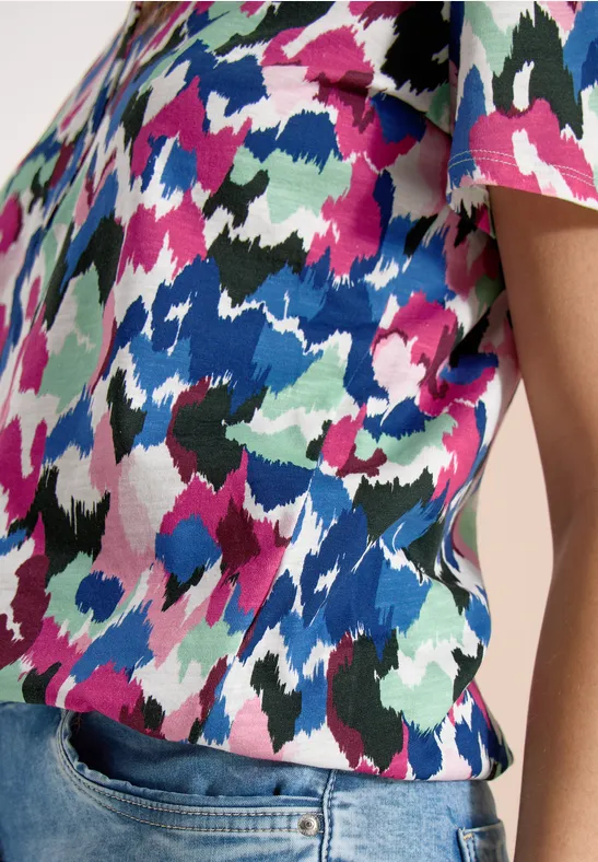 Person trägt kurzärmeliges Oberteil mit farbenfrohem, abstraktem Muster und eine Jeans.
