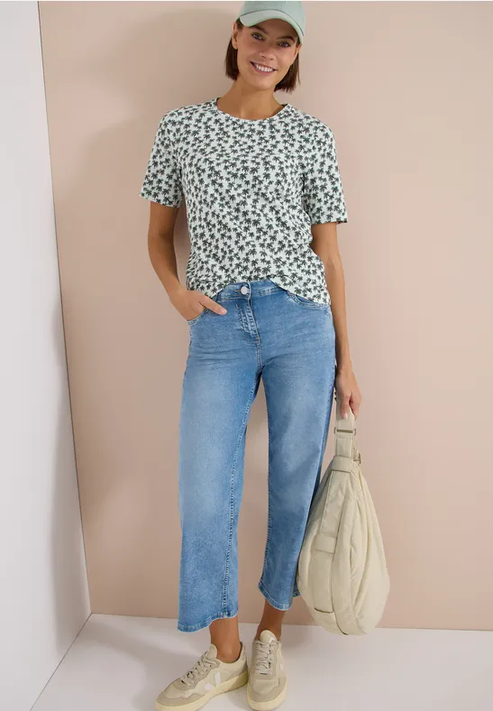 Person in gemustertem T-Shirt, geraden blauen Jeans, weißen Sneakern und mit einer Tasche.