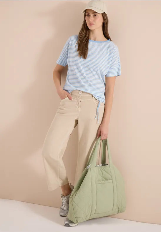 Frau lehnt vor heller Wand, trägt hellblaues T-Shirt, weite beige Hose und große grüne Tasche.