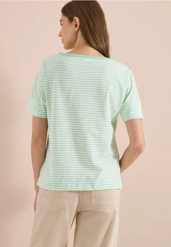Eine Frau steht mit dem Rücken zur Kamera, trägt ein grün-weiß gestreiftes T-Shirt und helle Hose