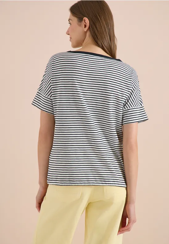 Person von hinten in schwarz-weiß gestreiftem T-Shirt und pastellgelber Hose vor hellem Hintergrund.