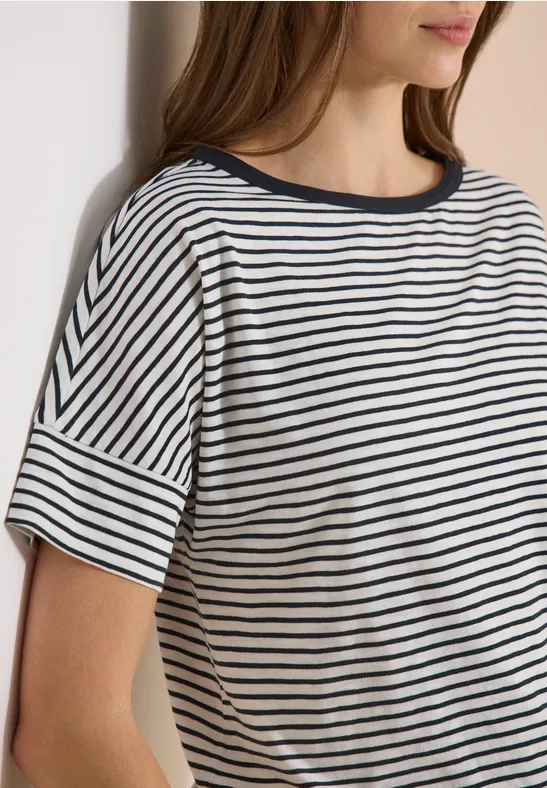 Frau trägt ein weiß-navy gestreiftes T-Shirt mit kurzen Ärmeln und lehnt an einer Wand.