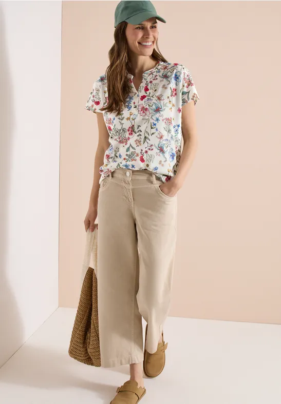 Vrouw in bloemen-T-shirt, lichte culottes, pet en rieten tas voor neutrale achtergrond.