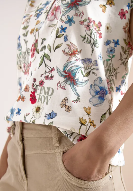 Witte top met kleurrijk bloemenpatroon, beige broek, hand steekt in de zak.