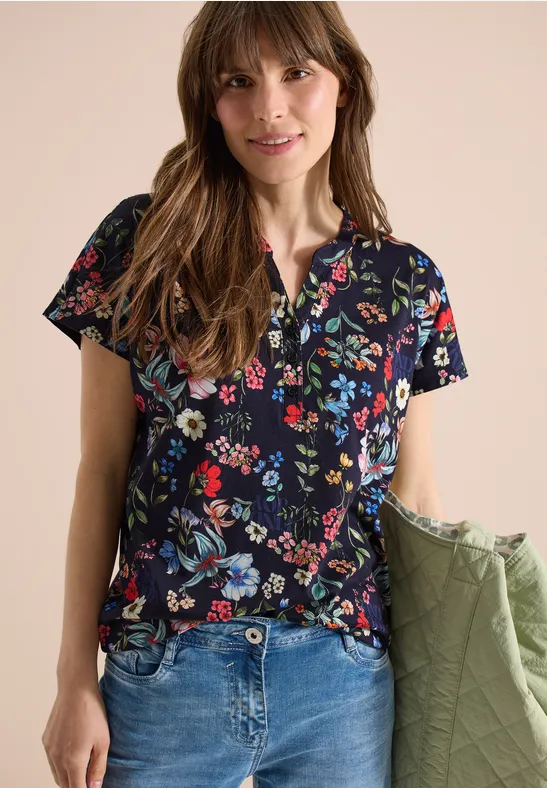 Vrouw draagt donkerblauw shirt met korte mouwen en bonte bloemenprint, een blauwe jeans en houdt een groene jas.