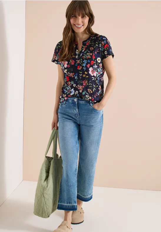 Vrouw in bloemen top, wijdvallende spijkerbroek, beige schoenen en groene tas tegen neutrale achtergrond.