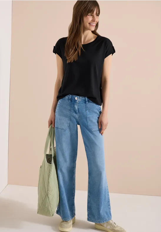 Frau in schwarzem T-Shirt, blauen weiten Jeans und grüner Tragetasche vor hellem Hintergrund.