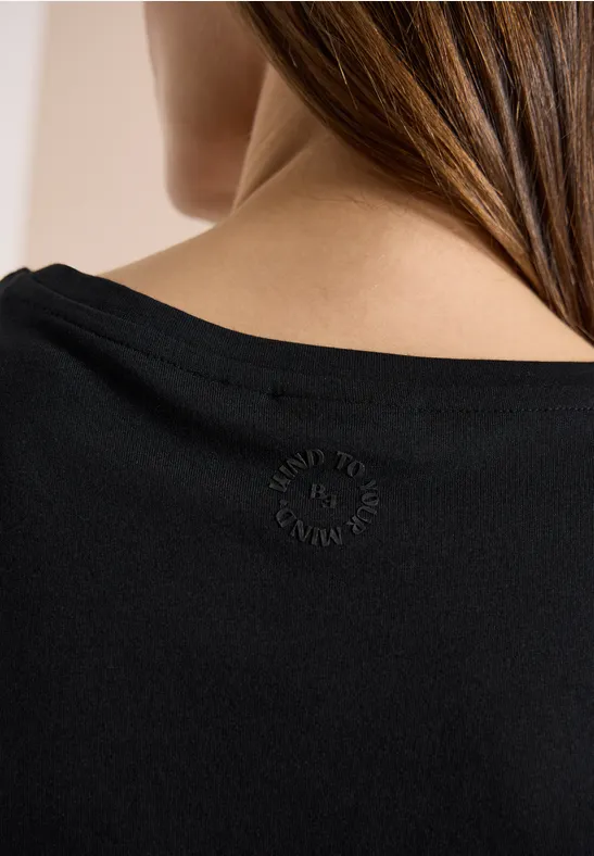 Rückansicht einer Frau mit langen Haaren in einem schwarzen Top, am Nacken ein kleines rundes Emblem.