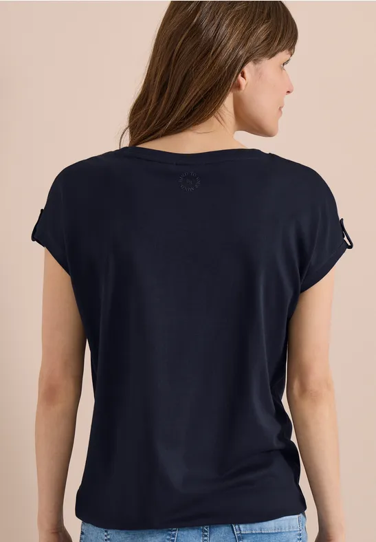 Vue de dos d'une femme en t-shirt bleu marine à manches retroussées sur fond beige.