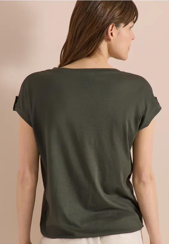 Rückansicht einer Frau mit halblangen braunen Haaren, trägt ein legeres dunkelgrünes T-Shirt vor beigem Hintergrund.