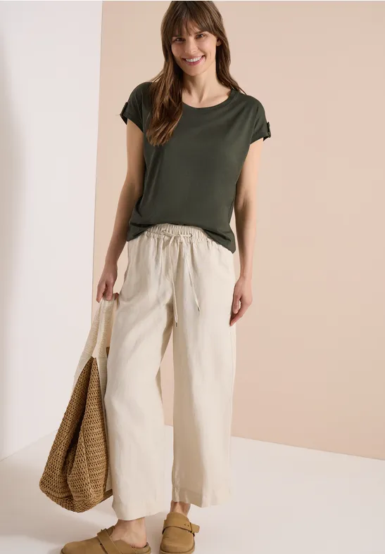 Frau in dunkelgrünem T-Shirt, cremeweißer weiter Hose, Basttasche und sandfarbenen Schuhen vor neutralem Hintergrund.