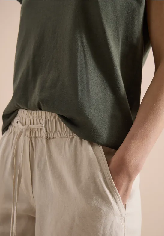 Grünes ärmelloses Top mit beiger Hose, Hand entspannt in Hosentasche.