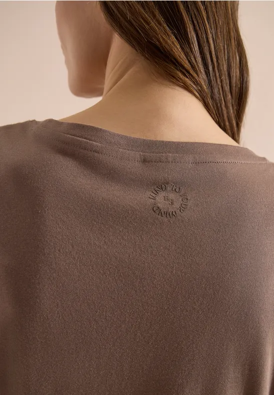 Vue de dos d'une femme portant un haut marron, cheveux tombant sur l'épaule, logo discret dans la nuque.