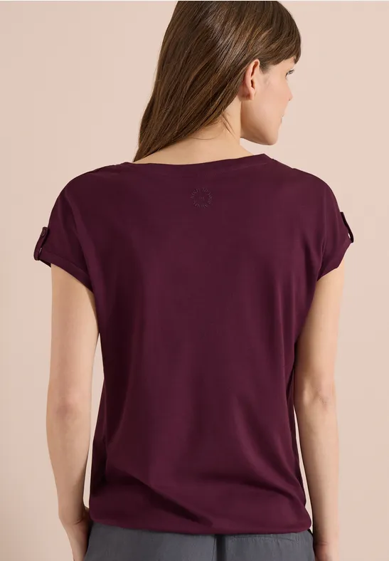 Femme de dos portant un t-shirt violet ample à manches courtes, fond beige.