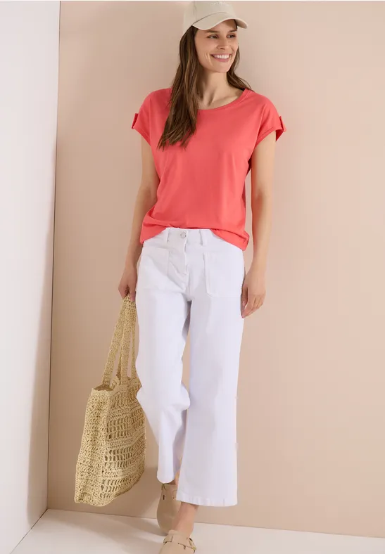 Frau in korallfarbenem T-Shirt, weißer Hose im Weit-Schnitt und geflochtenem Shopper vor hellem Hintergrund.