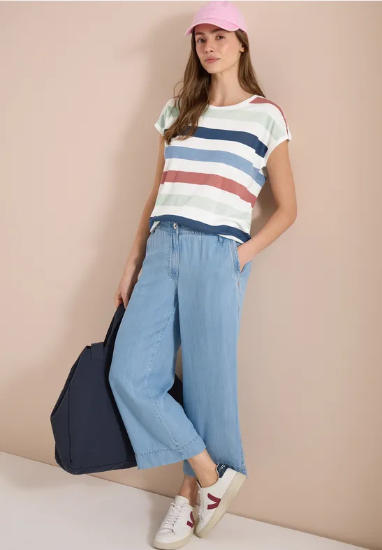 Vrouw in gestreept shirt, lichtblauwe culottes, witte sneakers, donkerblauwe tas, voor beige muur.