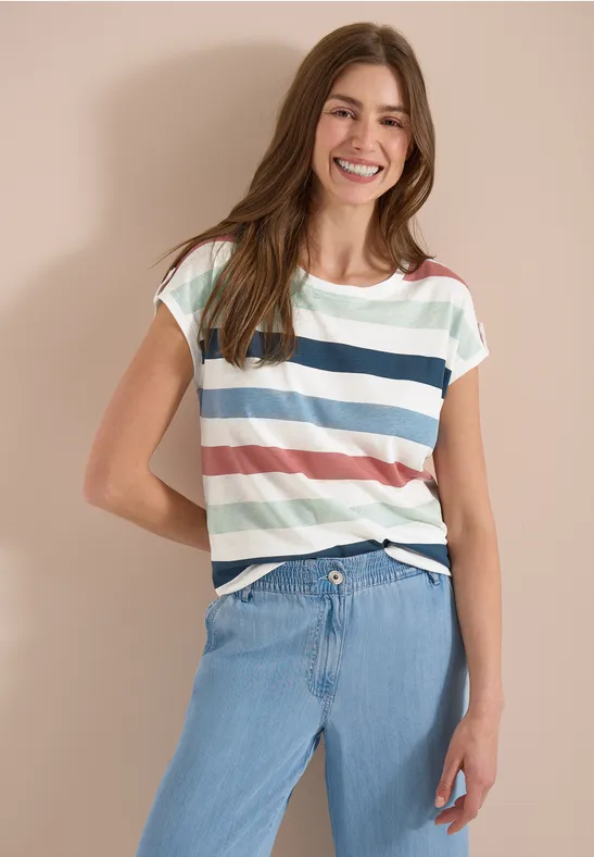 Een vrouw poseert voor een neutrale achtergrond, met een gestreept T-shirt en lichte spijkerbroek.