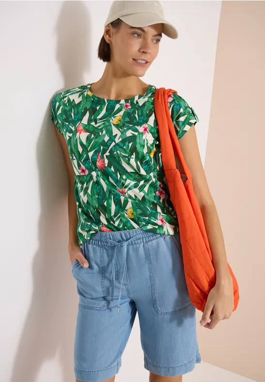 Frau in tropischem Print-Shirt, Jeansshorts, orangefarbener Stofftasche und Baseballcap.