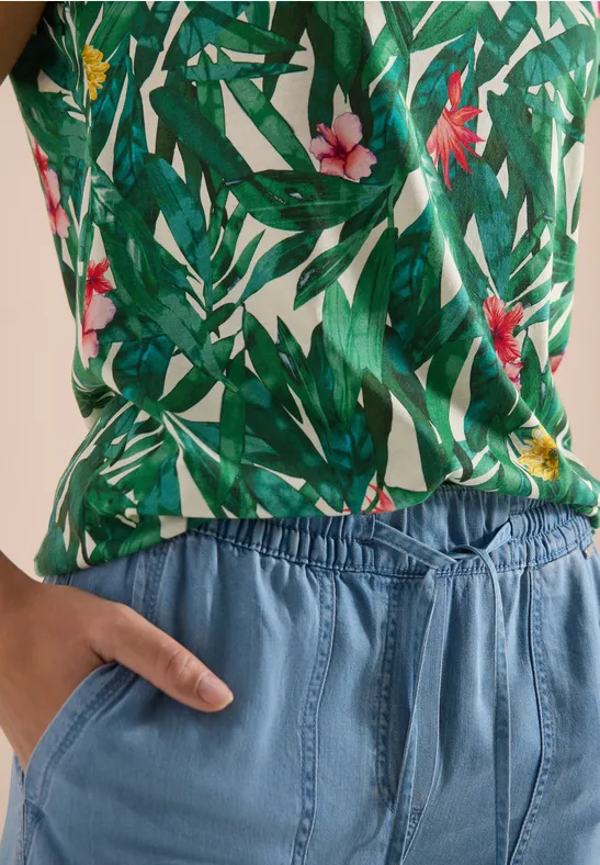 Person trägt grünes Top mit Blatt- und Blumenmuster und blaue Hose mit Gummizug, Hand in Tasche.