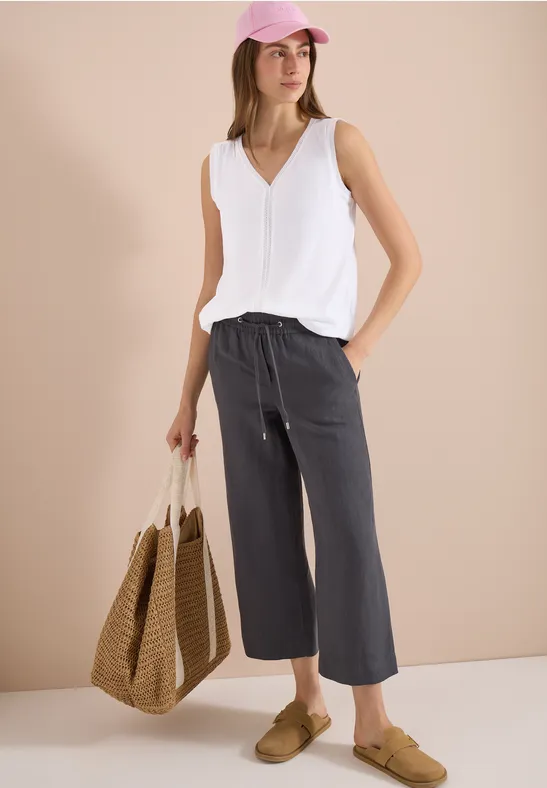 Model met witte V-hals top, losse grijze driekwartbroek, rieten tas, beige instappers en roze pet.