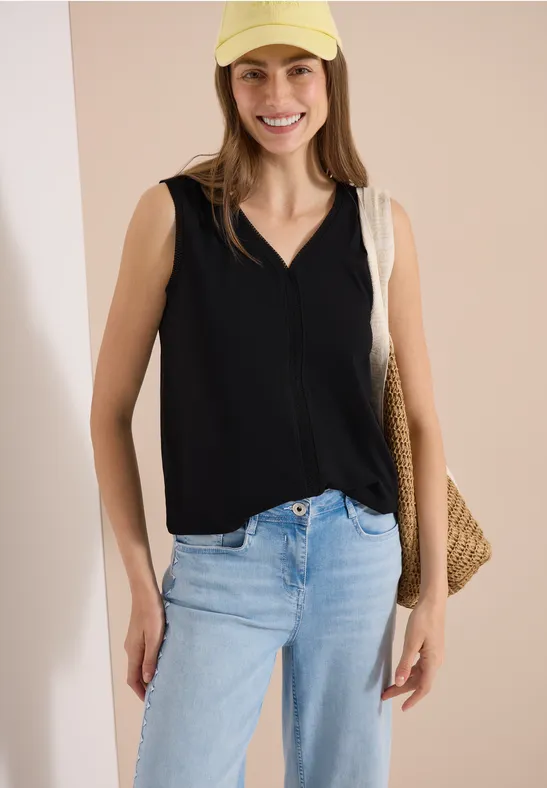 Vrouw draagt zwarte mouwloze top, lichte spijkerbroek en gevlochten strooien tas voor lichte achtergrond.