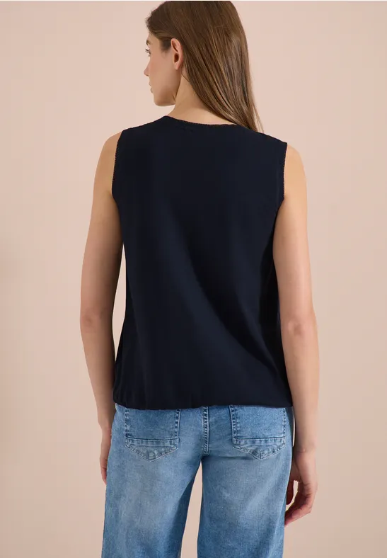 Achteraanzicht van een vrouw in een mouwloos donkerblauw topje en lichtblauwe spijkerbroek tegen neutrale achtergrond.