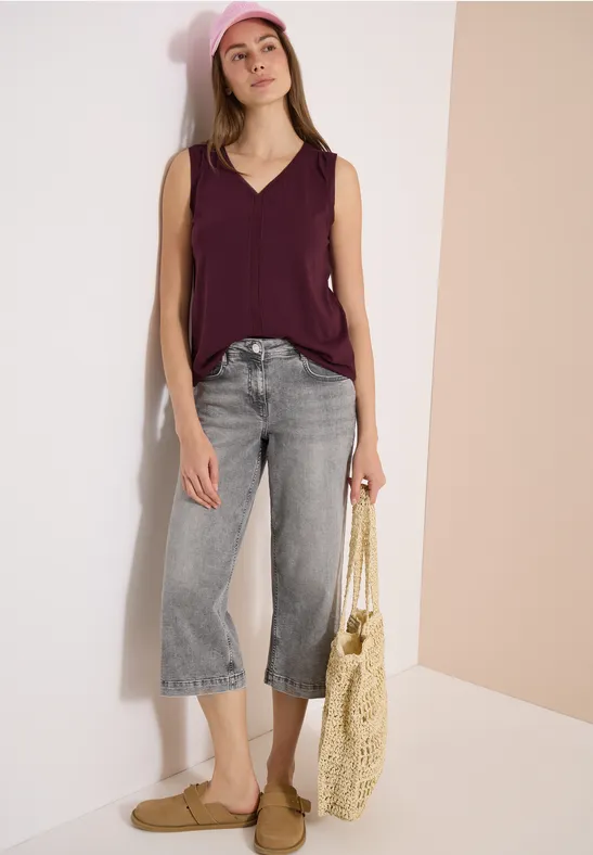 Frau trägt lila ärmelloses Top, graue weite 7/8-Jeans, Bastkorb und flache Sandalen vor hellem Hintergrund.