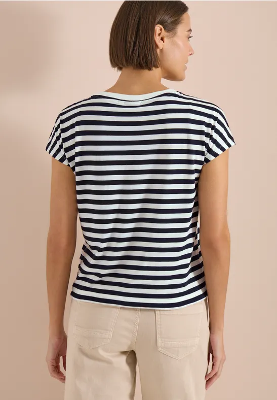 Une femme de dos porte un tee-shirt rayé noir et blanc à manches courtes et un pantalon clair.