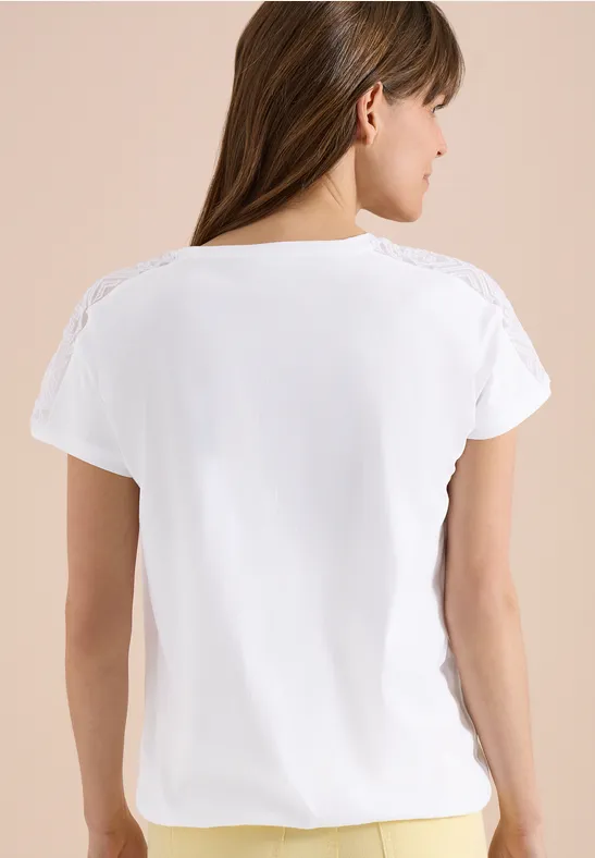 Eine Frau steht mit dem Rücken zur Kamera und trägt ein weißes T-Shirt mit Spitzenbesatz auf beigem Hintergrund.