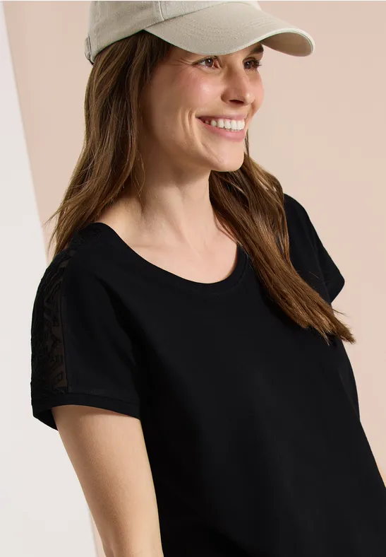 Eine Frau mit schwarzem T-Shirt und weißer Cap vor hellem Hintergrund, Gesicht unkenntlich.