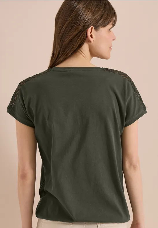 Femme de dos aux cheveux longs bruns, portant un T-shirt vert foncé uni devant un fond neutre.