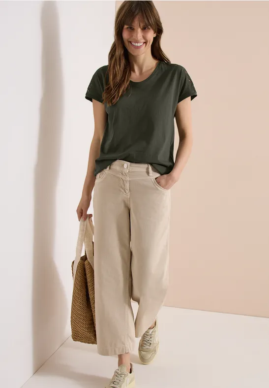 Femme en t-shirt kaki, pantalon ample beige et baskets, portant un sac en paille.