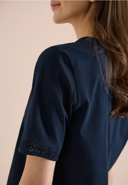 Vrouw in donkerblauw T-shirt met kanten detail aan de mouw, zijaanzicht van achteren.