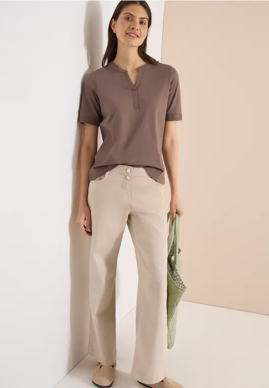 Vrouw in bruin T-shirt met korte mouwen en wijde beige broek, voor een lichte achtergrond, met een groene netzak.