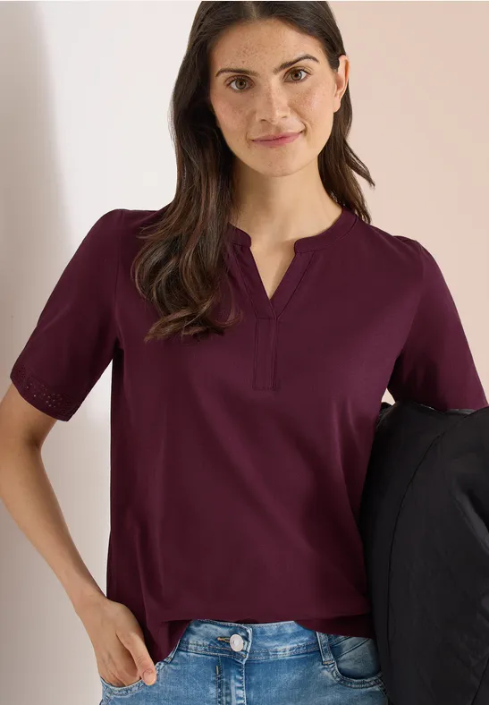 Een vrouw in een bordeauxrode blouse en spijkerbroek houdt een zwart jasje, gezicht onscherp.