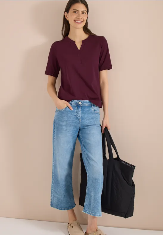 Vrouw met een bordeauxrood t-shirt, wijde cropped jeans en een zwarte tas tegen een beige achtergrond.