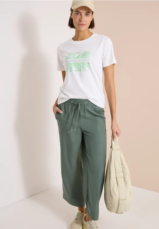 Vrouw in wit T-shirt met groen print, losse groene broek en houdt een lichte tas.