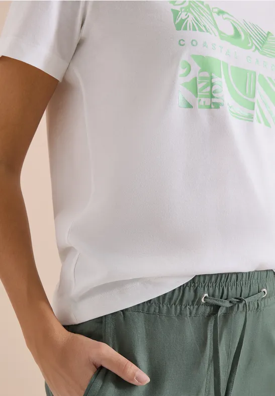 Een persoon draagt een wit T-shirt met groene print en een groene broek met koord, hand in zak.