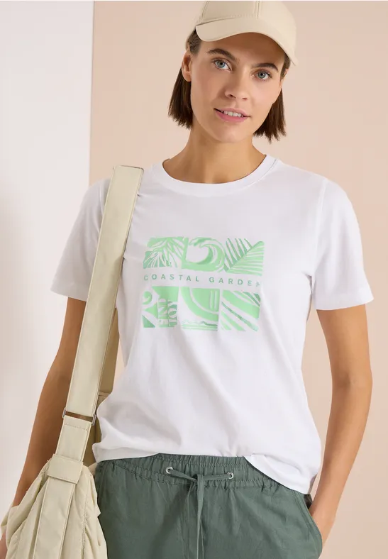 Persoon draagt wit T-shirt met groene print, groene broek, pet en lichte schoudertas.