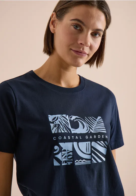 Persoon draagt donkerblauw t-shirt met 'COASTAL GARDEN' tekst en abstracte zee- en plantenpatronen.